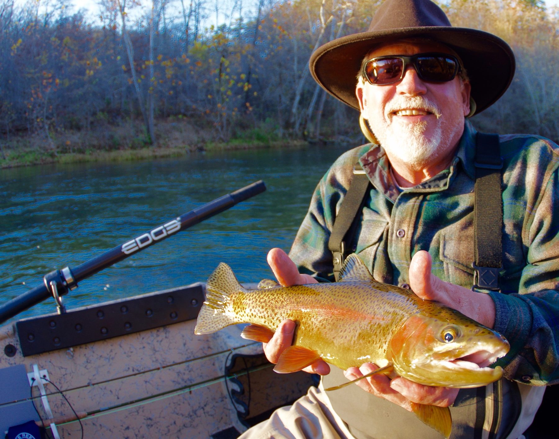 An angler in a felt hat and sunglasses holds a golden, spotted trout over the side of a boat