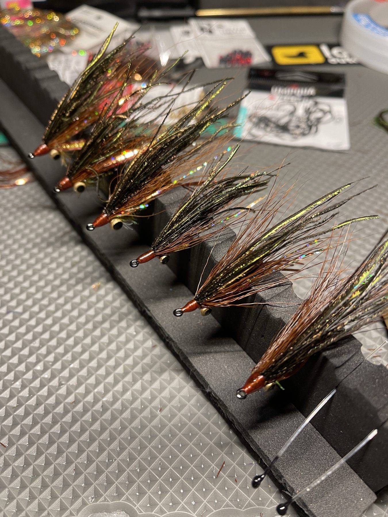 A row of custom hand-tied flies with peacock herl, copper hackle and bead-chain eyes on a tying foam block