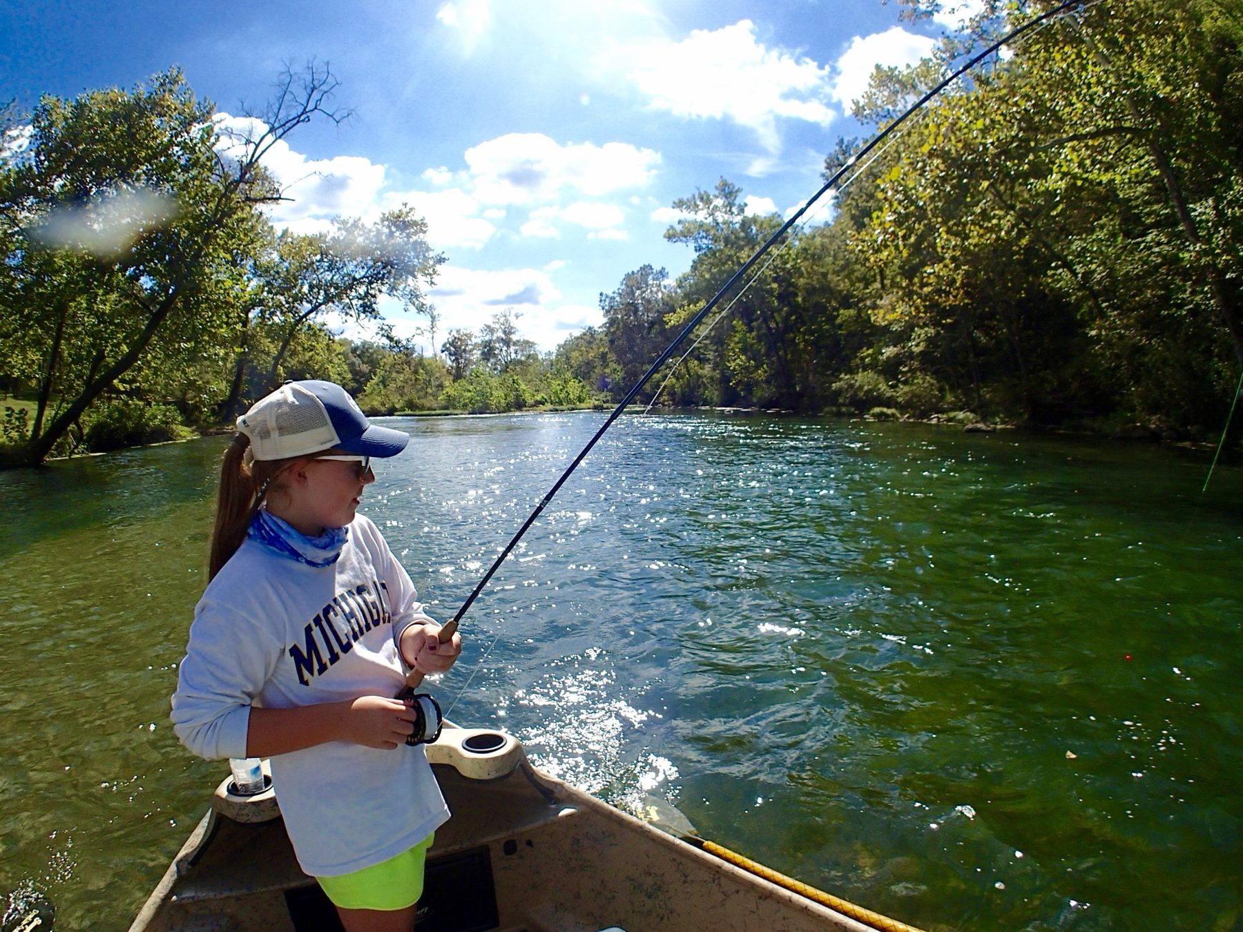 A young angler leans back against a bent fly rod on a sunlit green river