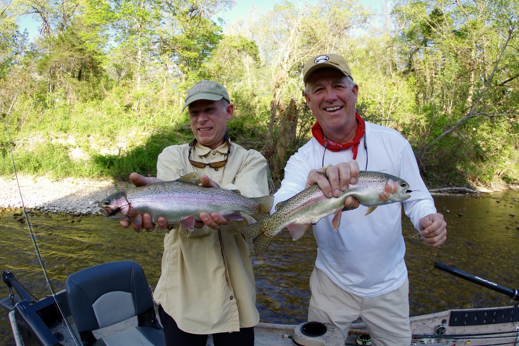 Two smiling anglers each holding a rainbow trout beside a drift boat on a gravel bar