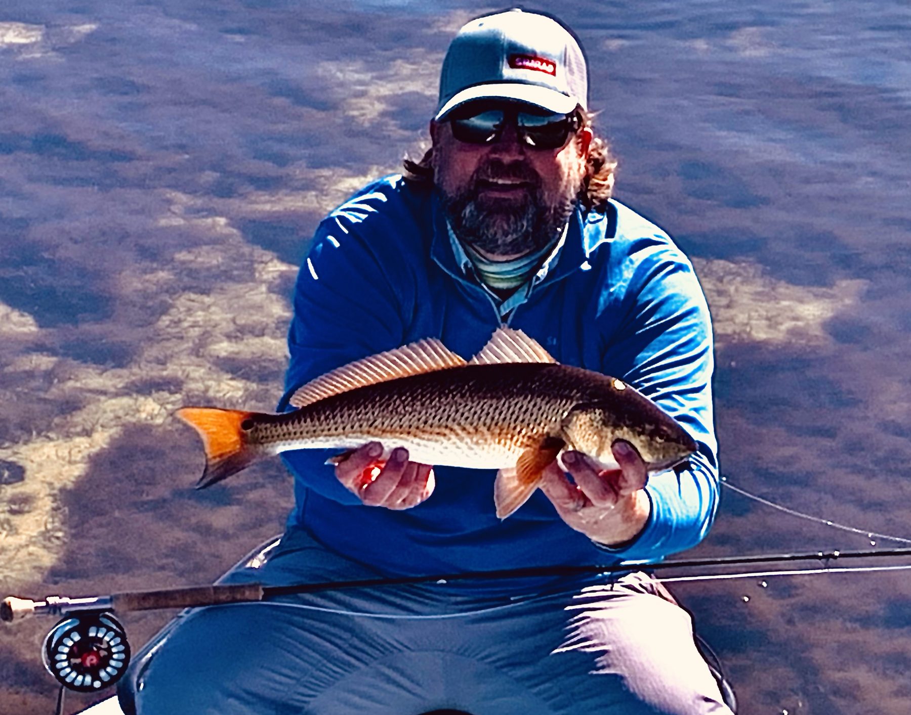An angler holds a redfish with a copper tail over clear shallow water, a fly rod across his lap