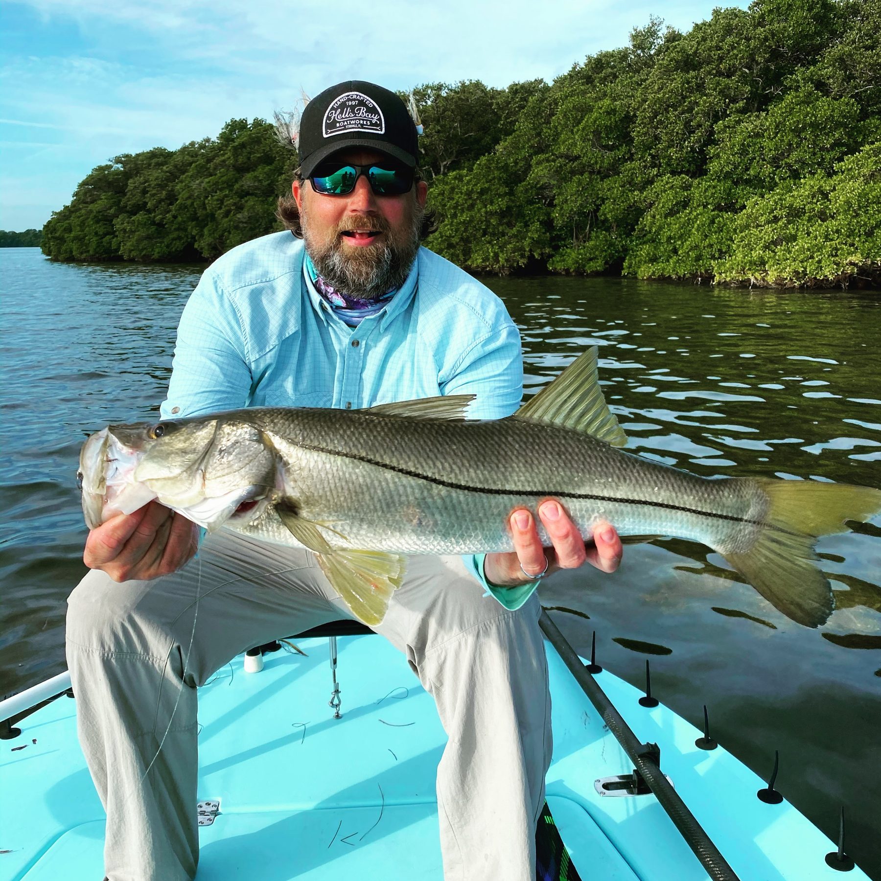 An angler in a bearded smile holds a snook from the bow of a flats boat beside a mangrove shoreline