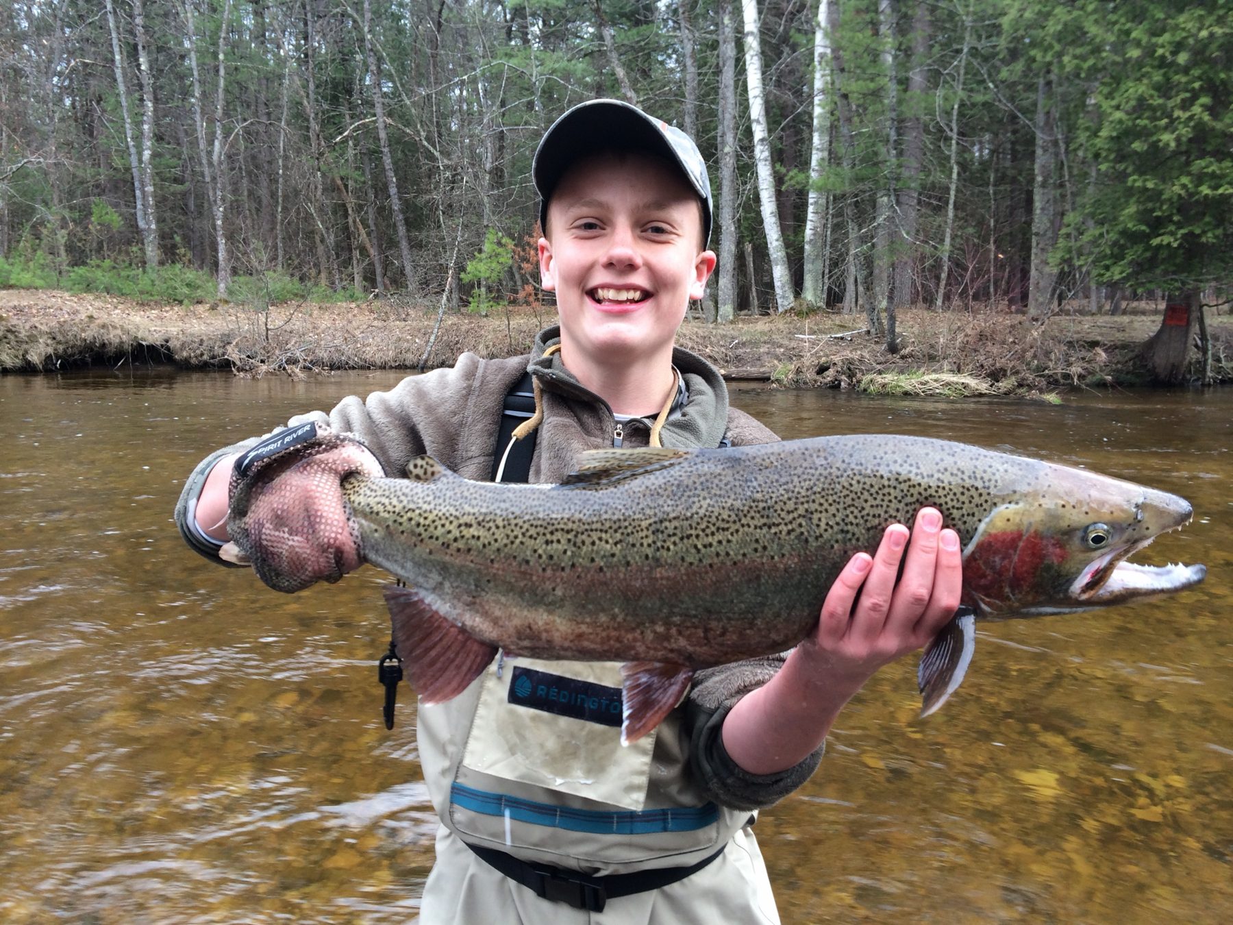 A smiling young angler in waders holds a large steelhead in a tannin-stained river