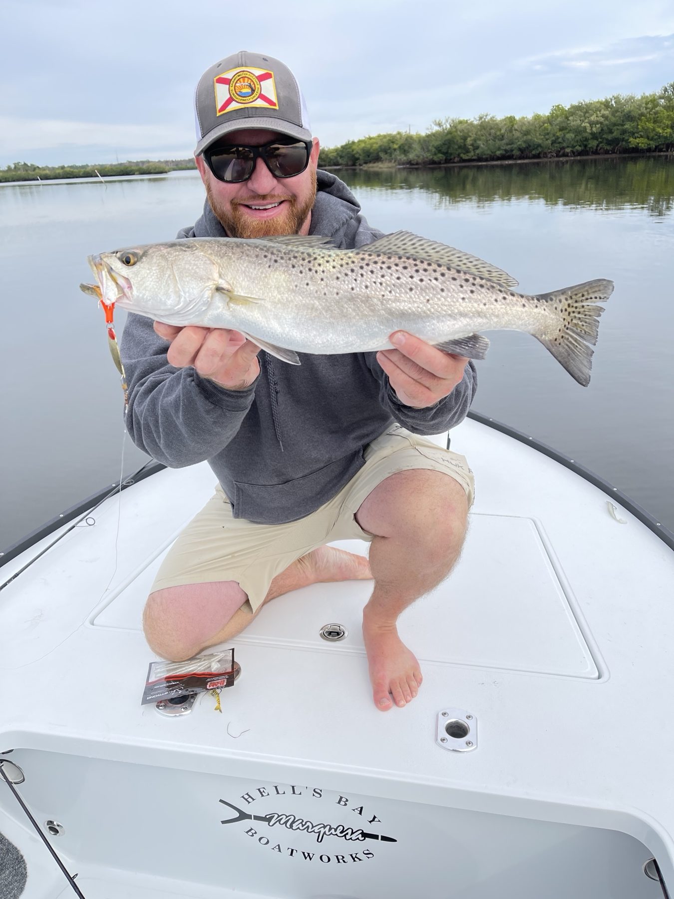 An angler in a ball cap kneels on the bow of a skiff holding a speckled trout on calm flats water